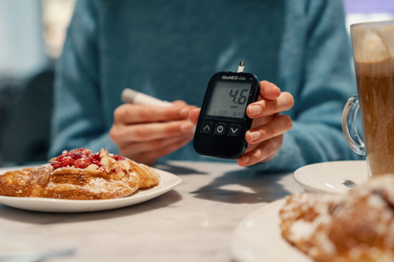 A person checks their blood sugar level using a glucometer while sitting at a table with a plate of pastry and a cup of coffee. The glucometer reads 4.6 mmol/L, indicating a normal blood sugar level. The image shows a casual setting where the individual is measuring their blood sugar before enjoying a snack.