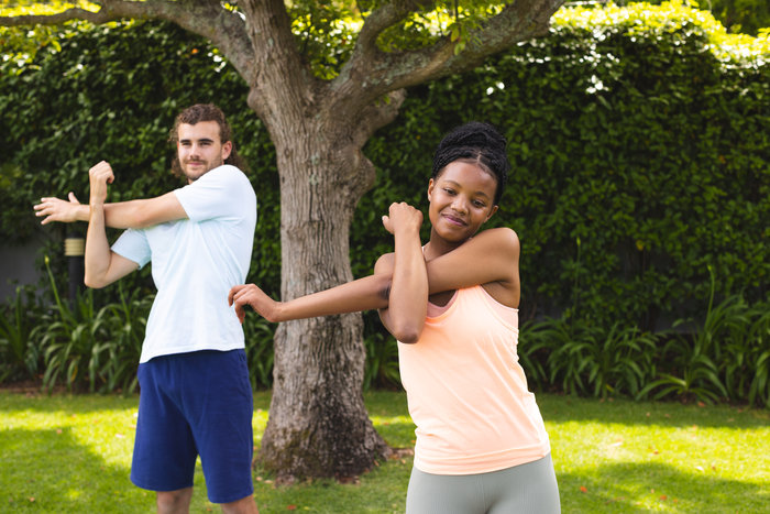 A young African American woman and a man stretch together outdoors in a park. They are both dressed in workout attire, with the woman smiling as she stretches her arm across her body. The man is stretching behind him. The background shows lush greenery and a tree, highlighting an active and healthy lifestyle.