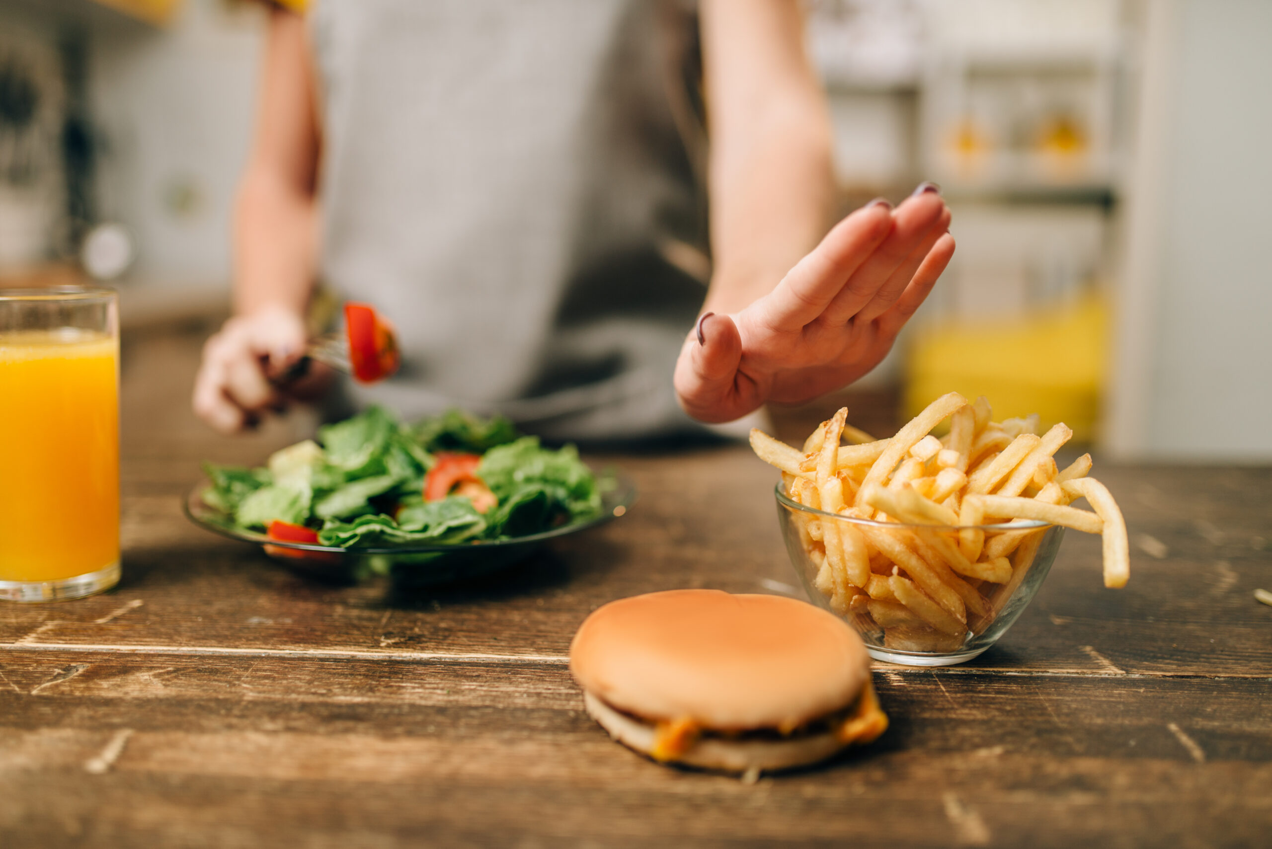 A woman is choosing healthy food over junk food. She is holding a fork with a piece of tomato and a salad in her left hand while using her right hand to gesture a "stop" towards a bowl of French fries and a burger on the table in front of her. A glass of orange juice is also visible on the table. The image conveys a decision to opt for healthier food choices.