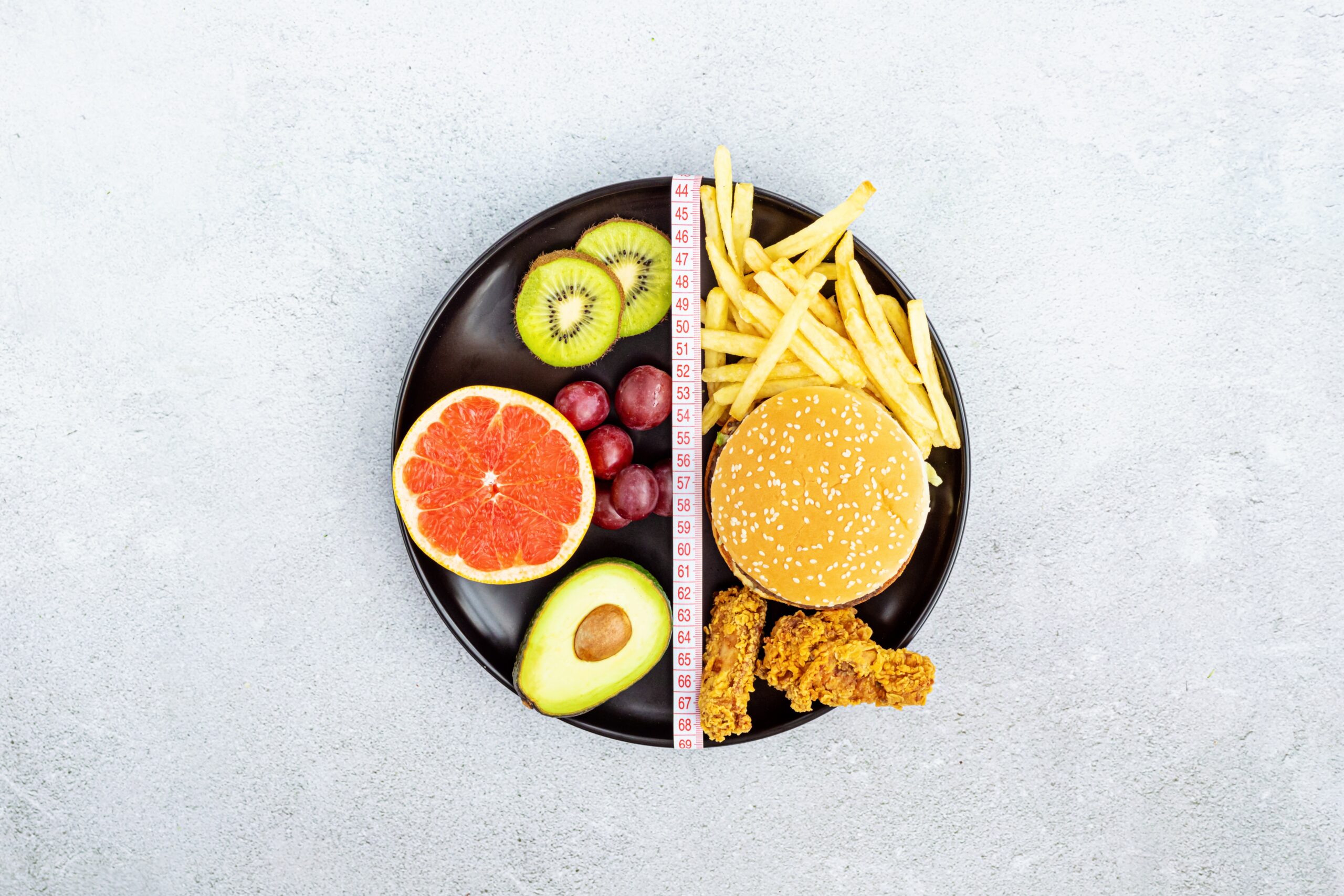 A plate divided into two halves: the left side features healthy foods like grapefruit, kiwi, grapes, and avocado, while the right side has unhealthy options like a burger, French fries, and fried chicken, with a measuring tape separating them.