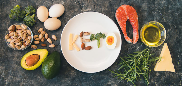 The photo displays a variety of keto-friendly foods arranged creatively to spell "Keto" on a plate. The ingredients include eggs, cheese, salmon, avocado, broccoli, almonds, pistachios, olive oil, and fresh herbs, emphasizing the healthy fats and proteins central to the Keto diet.