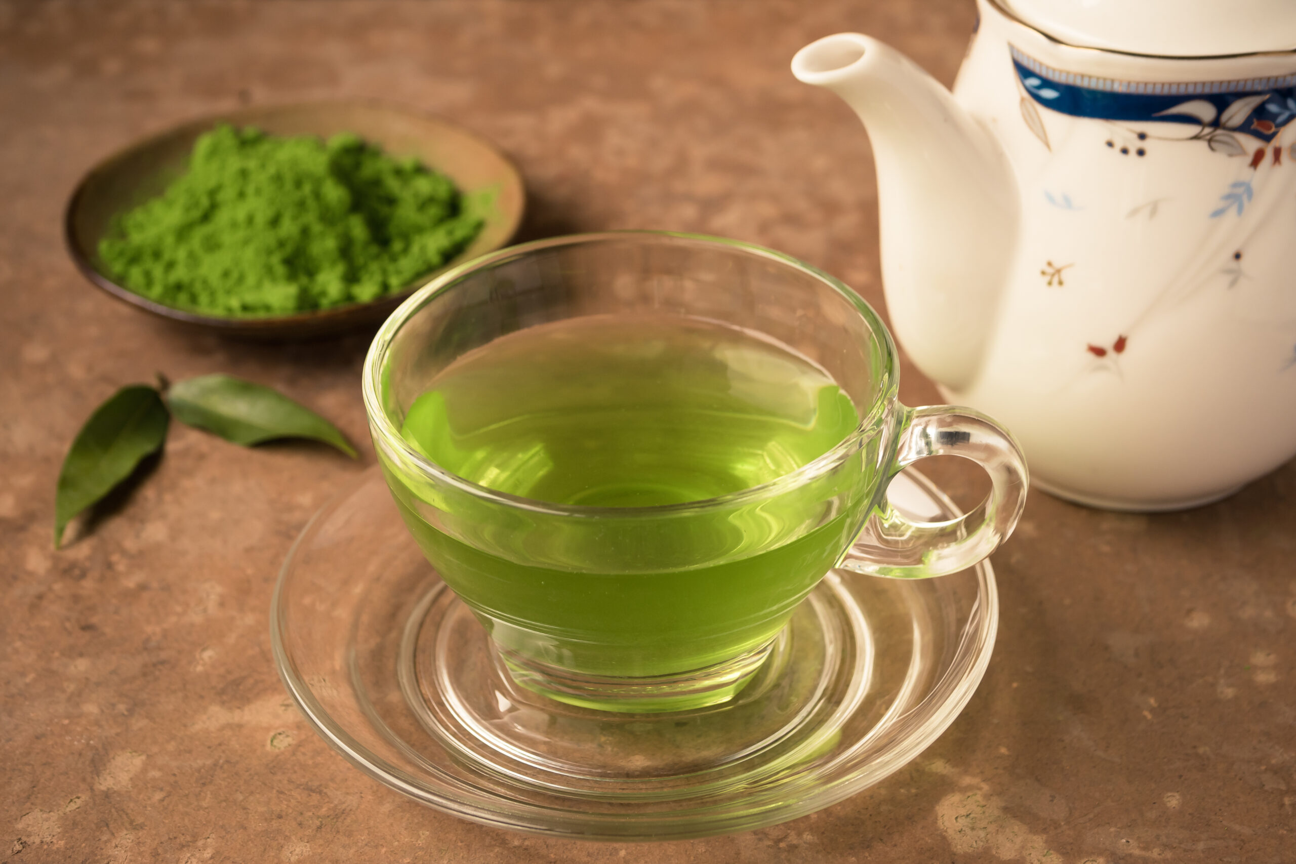A clear glass cup of green tea sits on a saucer, with matcha powder and a teapot in the background.