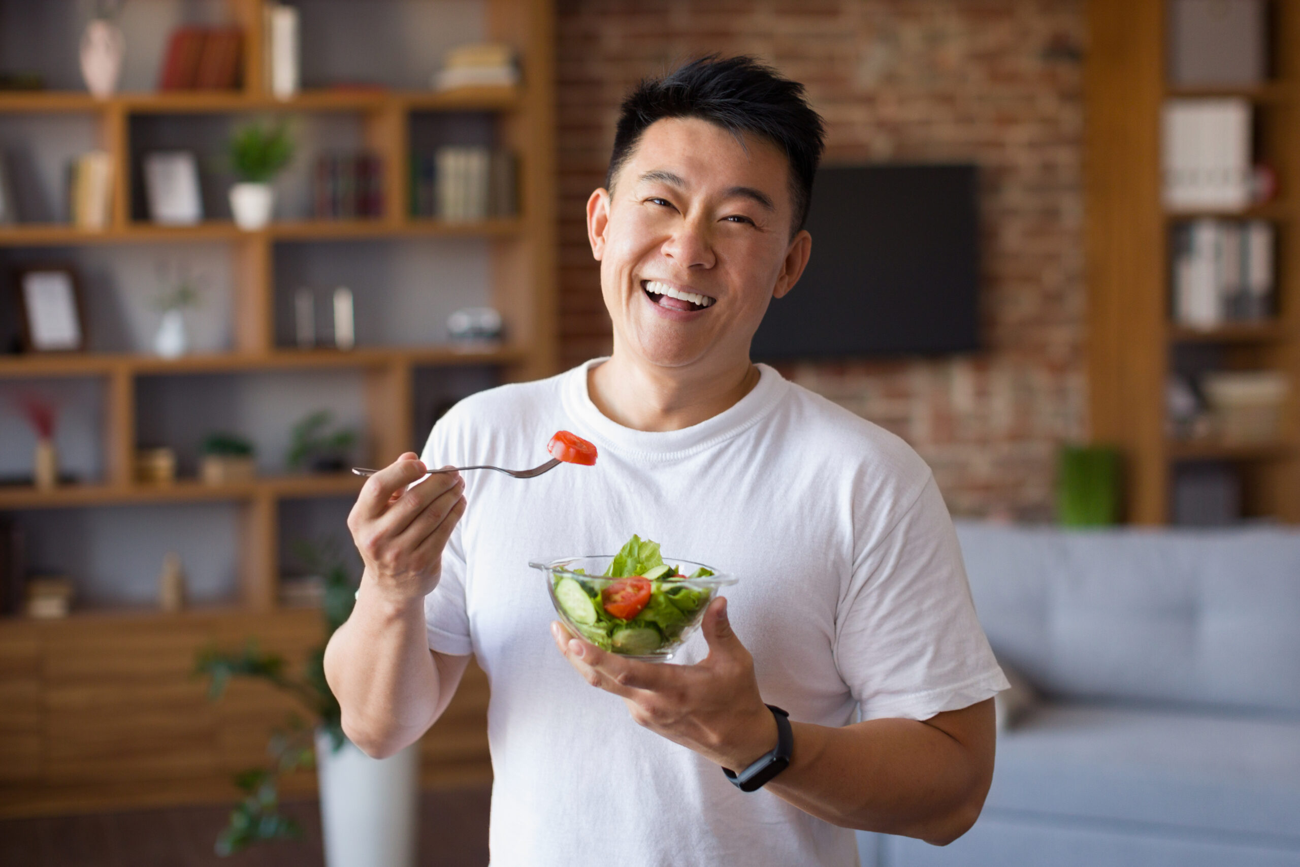 A smiling man enjoying a fresh salad in a relaxed, modern home setting.