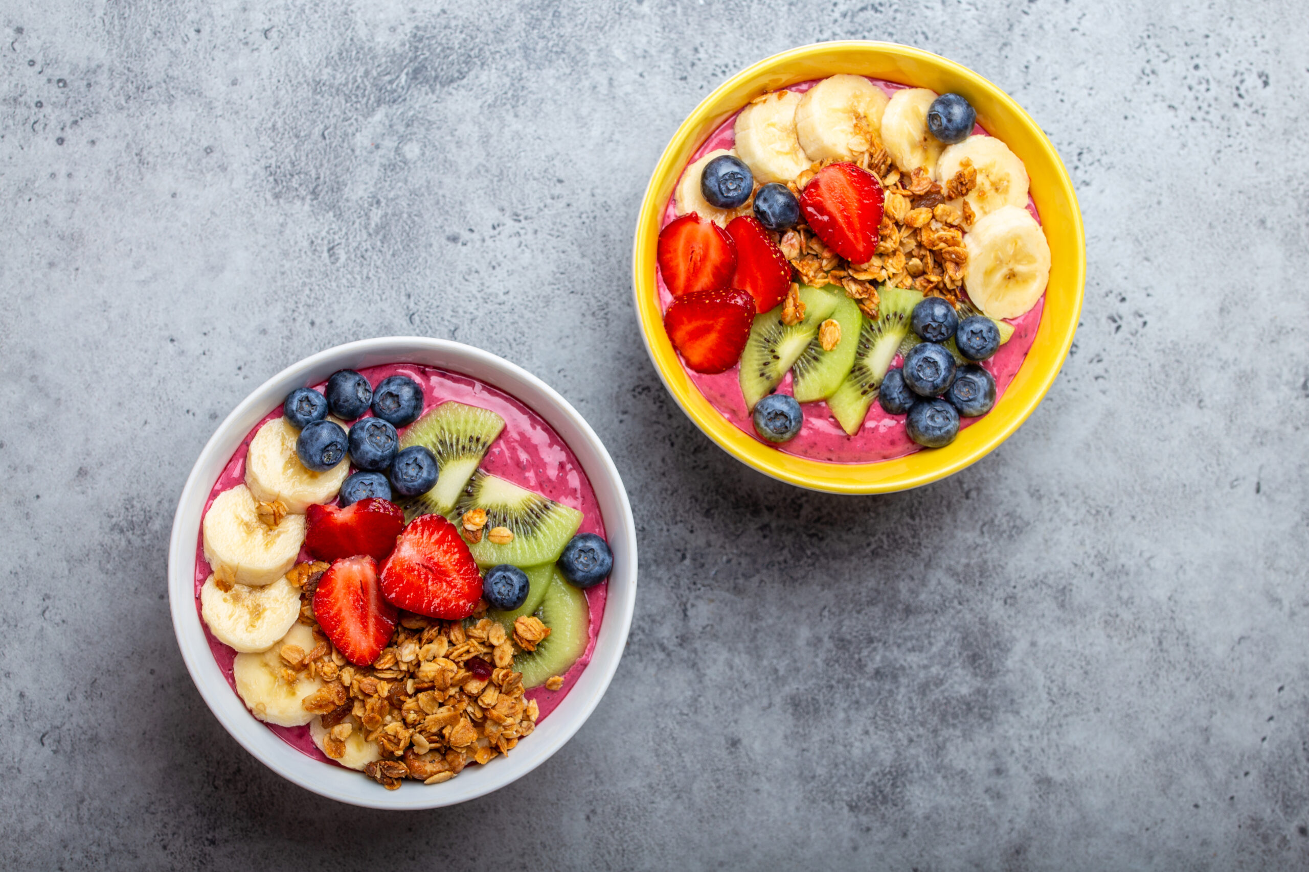 Smoothie Bowl: Two colorful smoothie bowls topped with fresh fruit such as banana, strawberries, kiwi, and blueberries, sprinkled with granola. 