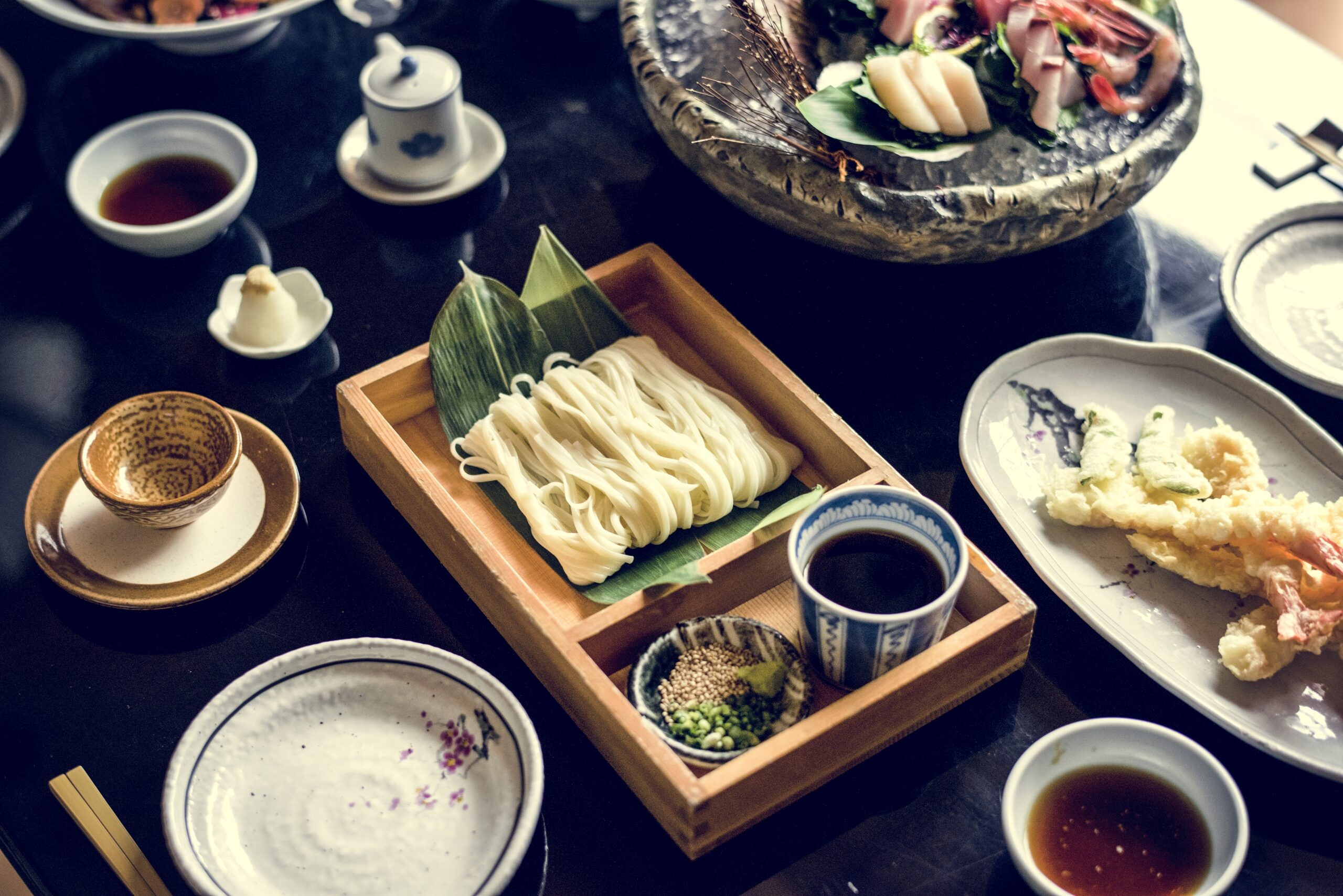 A Japanese meal set with cold noodles, dipping sauce, and tempura served on a dark table.