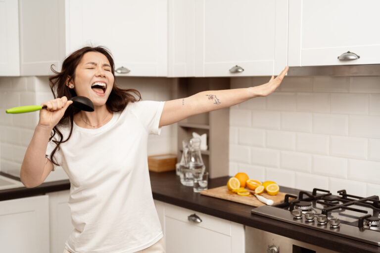 A woman joyfully singing in her kitchen while holding a green spatula as if it were a microphone. She is standing in front of a white kitchen cabinet, with her arm extended and a big smile on her face. There are slices of oranges on the kitchen counter, and a modern stove is visible in the background.