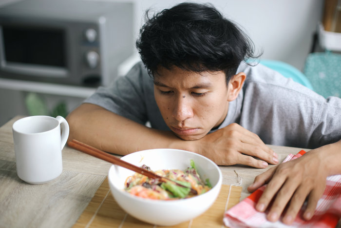 The image shows a man sitting at a table, looking disinterested or frustrated while staring at a bowl of vegetables. He appears unenthusiastic about eating the meal, with his face resting on his hand.