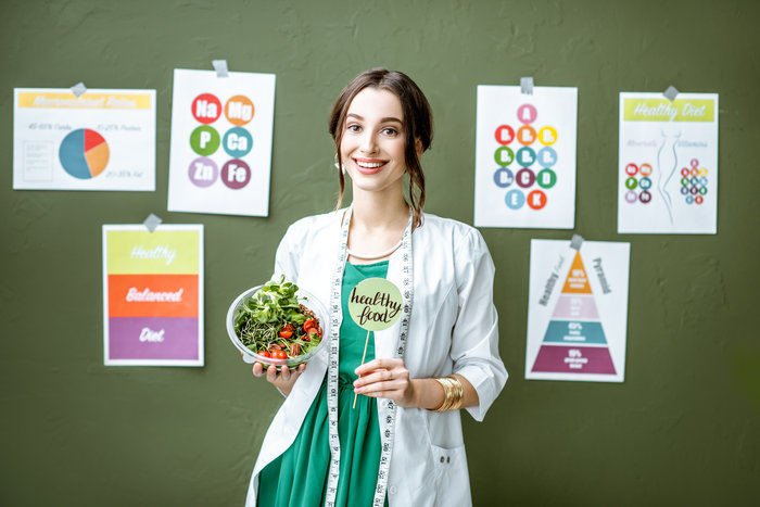 Portrait of a woman dietitian in medical gown standing with salad on the green wall background with drawings on a topic of healthy food indoors
