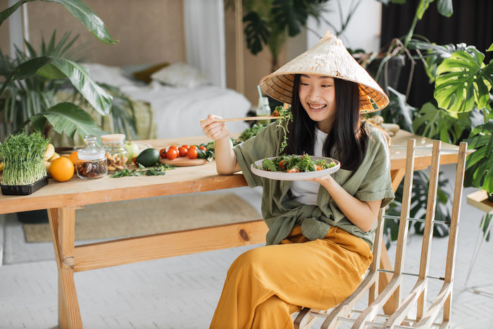 A smiling woman in a cozy, plant-filled room enjoys a fresh salad while wearing a traditional conical hat.