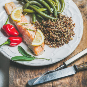 A close-up of a plate featuring roasted salmon garnished with lemon slices, served with quinoa, green beans, and red chili peppers, creating a colorful and balanced meal.