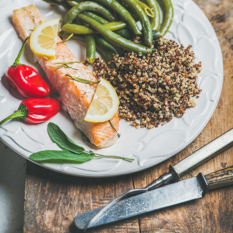 A close-up of a plate featuring roasted salmon garnished with lemon slices, served with quinoa, green beans, and red chili peppers, creating a colorful and balanced meal.
