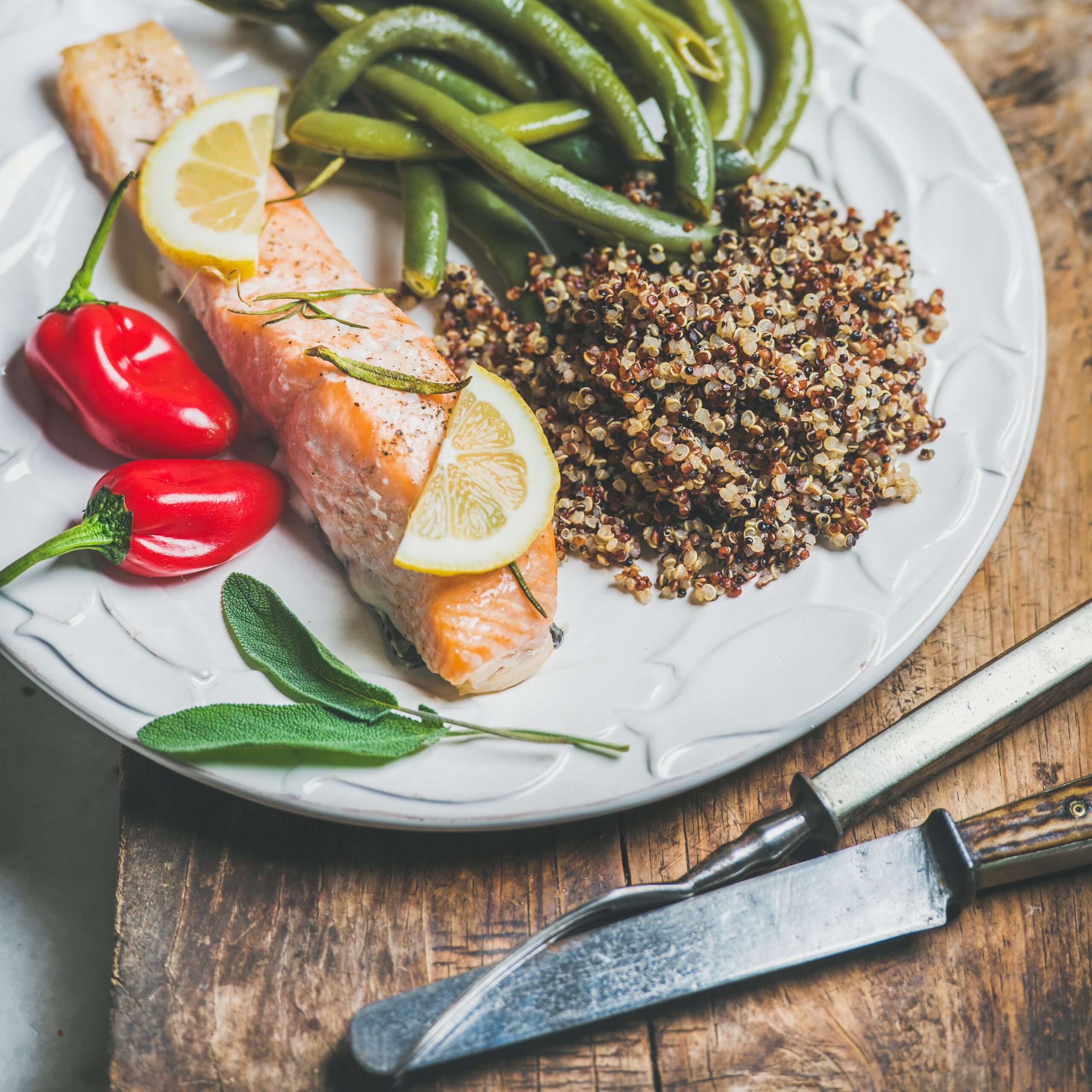 A close-up of a plate featuring roasted salmon garnished with lemon slices, served with quinoa, green beans, and red chili peppers, creating a colorful and balanced meal.