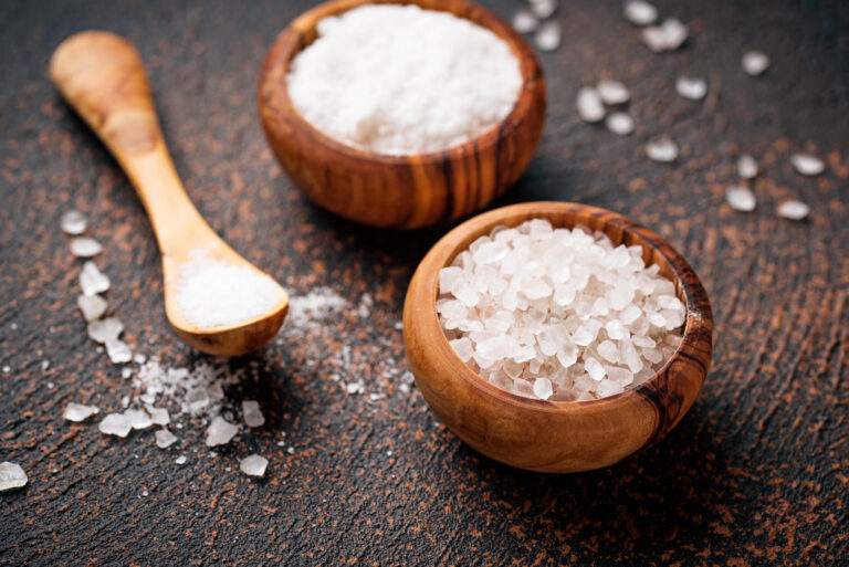 Two wooden bowls filled with different types of salt — one with coarse salt crystals and the other with fine white salt — accompanied by a wooden spoon, all arranged on a rustic dark surface.
